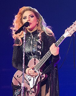 Lady Gaga standing behind a microphone stand with a pink guitar in her hands, wearing black leather fringe