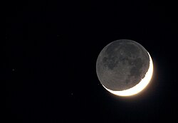 An overexposed photograph of a crescent Moon reveals earthshine and stars.