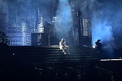 An extreme wide shot of the Weeknd performing with a durag in white-colored fashion as he sits down and performs on the staircase of the concert's setup.