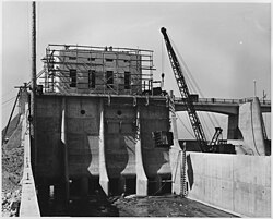 Looking upstream in the outlet channel. Form work has been stripped from the outlet control tower and service bridge and one of the outlet control slide gate frames is being lowered into position for installation.