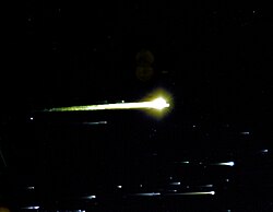 White streaks of light, with bright spots on the right side of them, fill the bottom of the frame. A larger yellow-tinted sphere with a streak is in the center of the frame. The background is black space.
