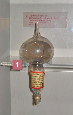 The inside of a museum exhibit shows a heavily used light-bulb with smoky glass. There is a handwritten inscription on a label wrapped around the stem of the bulb.
