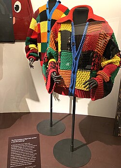A knitted cardigan with yellow, red, orange, green and black squares, displayed at a museum
