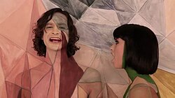 Upper body shot of a man (left) and a woman turned towards him. The man is taller and has dark stragly hair. The woman has dark hair cut in a bob. Both have skin painted in coloured geometric designs. Both are singing. The backdrop has a similarly coloured design closely matching the design on the two people.