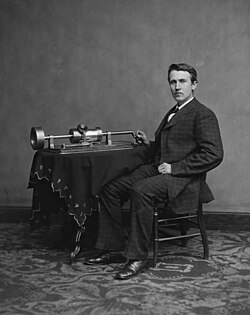 Edison as a young man, sharply dressed, seated over decorative carpet and facing slightly towards the right with his face towards the camera. His right hand positioned to start turning his phonograph.