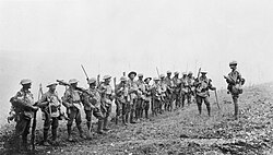 A line of soldiers in battle equipment face another soldier who is addressing them on a gentle slope. Behind them smoke or fog obscures the rest of the terrain.