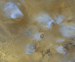 Orographic water ice clouds hover over the volcanic peaks of the central Tharsis region in this color image mosaic from Mars Global Surveyor. Olympus Mons dominates at upper left. At center are the three Tharsis Montes: Arsia Mons at bottom, Pavonis Mons at center, and Ascraeus Mons at top.