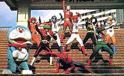 Protagonists of popular tokusatsu franchises mostly of the late 1970s (from back to front, left to right): Ultraman Joneus (Ultra Series), Battle Fever J (Super Sentai), Kamen Rider Stronger and Kamen Rider V3 (Kamen Rider Series), and Spider-Man. The photo also features manga character Doraemon on the far left.