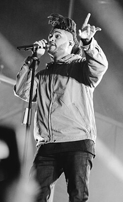 The Weeknd, sporting a Basquiat-like hairstyle, holds a microphone while his other hand points up as he performs.