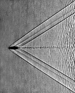 Shadowgram of shock waves from a supersonic bullet fired from a rifle. The shadowgraph optical technique reveals that the bullet is moving at about a Mach number of 1.9. Left- and right-running bow waves and tail waves stream back from the bullet and its turbulent wake is also visible. Patterns at the far right are from unburned gunpowder particles ejected by the rifle.