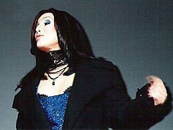 Cher performs in a glittering blue outfit with a black coat and layered necklaces, with sleek, straight dark hair.