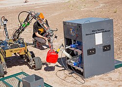 Rover next to a metal generator, holding a can of fuel. Both are on a vast desert