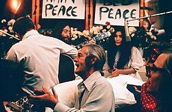 Lennon and Ono sit in front of flowers and placards bearing the word "peace". Lennon is only partly visible, and he holds an acoustic guitar. Ono wears a white dress, and there is a hanging microphone in front of her. In the foreground of the image are three men, one of them a guitarist facing away, and a woman.