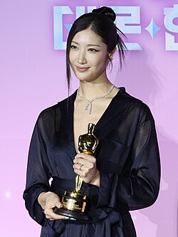Ejae from the waist up holding her Oscar trophy with both hands. She is looking off slightly left of center.