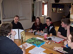 A group playing a tabletop role-playing game (RPG). The Games Master is at left using a cardboard screen to hide dice rolls from the players.