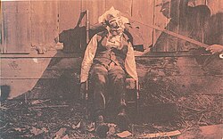 Body of a lynched Black male, propped up in a rocking chair for a photograph, c. 1900. Paint has been applied to his face, circular disks glued to his cheeks, cotton glued to his face and head, while a rod props up the victim's head.
