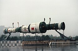 A full-scale model of a Salyut 7 space station and two docked spacecraft. On the left a Soyuz can be seen docked to the fore port, and on the right a Progress is docked at the aft port. The display is in front of one of the pavilions of the Exhibition of Soviet National Economic Achievement.