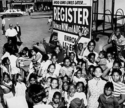 A group of African-American children gather around a sign and booth to register voters. Early 1960s.