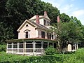 A rambling yellow two-story house, featuring a porch wrapping around the left side, three chimneys, and a square turret rising from the rear.