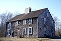 A traditional 2-story colonial-style house, with weathered cedar shake siding and roofing. A woman stands near the entrance.