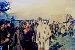 Australian officer casually dressed in only shorts holding a bow and other weapons while others look on