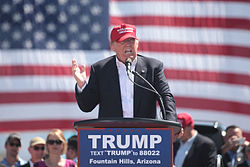 Trump speaking in front of an American flag behind a lectern, wearing a black business suit and a bright red "MAKE AMERICA GREAT AGAIN" baseball cap. The lectern sports a blue "TRUMP" sign.