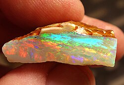 A small opal held side-on between someone's forefinger and thumb. A small sliver of brown rock is visible at the top, with a larger section of opal below it.