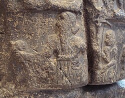 Sargon on his victory stele, with a royal hair bun, holding a mace and wearing a flounced royal coat on his left shoulder with a large belt (left), followed by an attendant holding a royal umbrella.[51][52] The name of Sargon in cuneiform ("King Sargon") appears faintly in front of his face.[51][53] Louvre Museum.