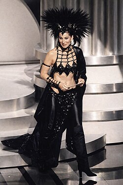Cher stands on the Academy Awards stage holding a presenter's envelope, wearing a black Bob Mackie ensemble featuring a feathered headdress, an open lattice-work top exposing her midriff, sequined pants, and a long cape.