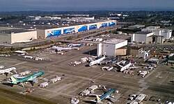 An aerial photograph of a large factory and an airport with several jetliners surrounded by equipment and ground vehicles.
