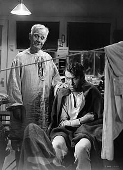 Travers stands behind a seated Stewart putting his hand on Stewart's shoulder