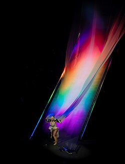 Gaga at her Mayhem Ball tour in 2025, with rainbow lights projected onto her long white cape[461]