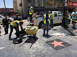 Construction workers in high-vis yellow vests work with trash bags and a wheelbarrow on the sidewalk around a recently poured terrazzo pad and unidentifiable star. Police officers in front of caution tape and pedestrians behind caution tape watch. Disney, Ghirardelli, and TMZ banners are visible across the street.