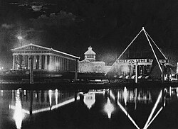 Greek architecture pictured at night in black in white. The foreground is occupied by a small body of water and the building in the middle and background have their perimeter outlined with string lights.