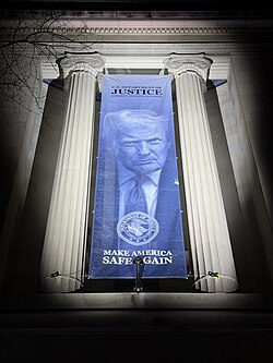 Nighttime photograph of a large vertical banner hanging between two neoclassical limestone columns. The banner shows Trump's official portrait, with "U.S. Department of Justice Robert F. Kennedy Building" above and "Make America Safe Again" below.
