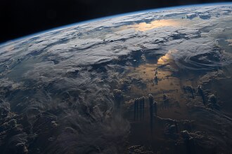 Earth from orbit, showing sunlight filtered into a reddish hue by the troposphere, while casting shadows from different cloud types. The stratosphere forms a thin band of blue scattered sunlight along the horizon.