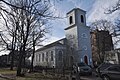 A gray church stands next to an old cemetery. The church has a tower but no steeple, and a circular drive in front.