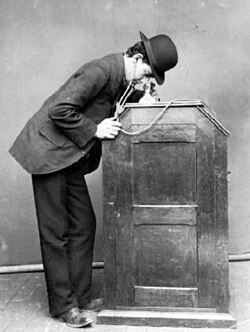The 1895 version of the Kinetophone in use, showing the earphones that lead to the cylinder phonograph within the cabinet.