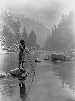 Photo of man standing on rock holding spear with spearpoint in the water