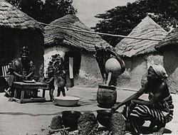 A housewife in Yendi, Kumasi, Ghana, pours water into a meal as her children play, 1957