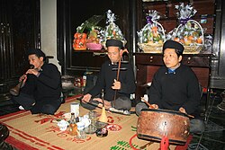 A trio of Vietnamese musicians performing together. The man on the far left plays kèn đám ma, the man in the middle plays the đàn nhị and the man on the right plays the trống chầu.