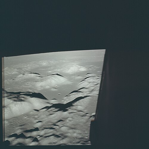 The valley of Taurus-Littrow as seen from the Lunar Module Challenger on the orbit before powered descent there. The Command and Service Module America can just be seen crossing the base of the 2.3 km high South Massif. Between the South and North Massifs, the valley is 7 km wide. Mare Serenitatis, the Sea of Serenity, is on the horizon.