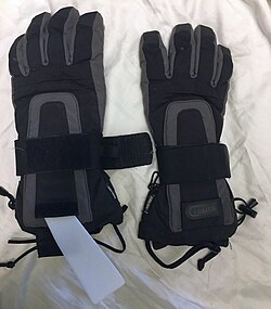 Gloves with an integrated plastic element, seen left partially pulled out, as a wrist guard. The element is pressed tightly to the wrist with a wide, all-around velcro-fastened strap, which can be seen in loose state on the left and tightened on the right glove. This stabilizes the wrist and thus avoids its damage when falling onto the hands.