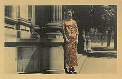 A handcoloured photo of a woman wearing traditional Chinese clothing standing next to the State Library of Victoria, c. 1920s