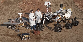 Two Jet Propulsion Laboratory engineers stand with three vehicles, providing a size comparison of three generations of Mars rovers. Front and center left is the flight spare for the first Mars rover, Sojourner, which landed on Mars in 1997 as part of the Mars Pathfinder Project. On the left is a Mars Exploration Rover (MER) test vehicle that is a working sibling to Spirit and Opportunity, which landed on Mars in 2004. On the right is a test rover for the Mars Science Laboratory, which landed as Curiosity on Mars in 2012. Sojourner is 65 cm (26 in) long. The Mars Exploration Rovers (MER) are 1.6 m (5 ft 3 in) long. Curiosity on the right is 3 m (9.8 ft) long.