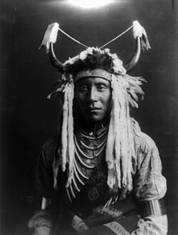 Head Carry, a Piegan man wearing a split horn headdress. Photographed by Edward S. Curtis, 1900.