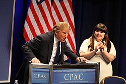 Trump, leaning heavily onto a lectern, with his mouth open mid-speech and a woman clapping next to him