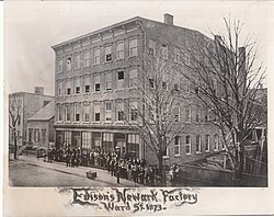 Group photo taken as a postcard of the employees in front of the factory at Edison's Newark Factory on Ward St in 1873. The trees are bare and the surrounding buildings are much smaller. Edison is visible peeking out of the fourth floor window. He wrote that he was too busy working to come downstairs.