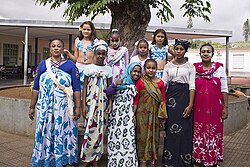 Mahoran women and girls pose together in a group. They are wearing the traditional colorful salouva of Mayotte, and some of them also have a matching kichali. Their arms are covered by cotton body garments. They are wearing makeup, but only the woman on the right and the young girls have facial designs drawn with msinzano
