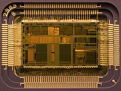 An integrated circuit (IC) as an example application in the field of microelectronics. The chip housing is opened to allow a view of the actual circuit. The golden connecting cables, which form the electrical wiring between the IC and the housing contacts, can be seen on the sides.